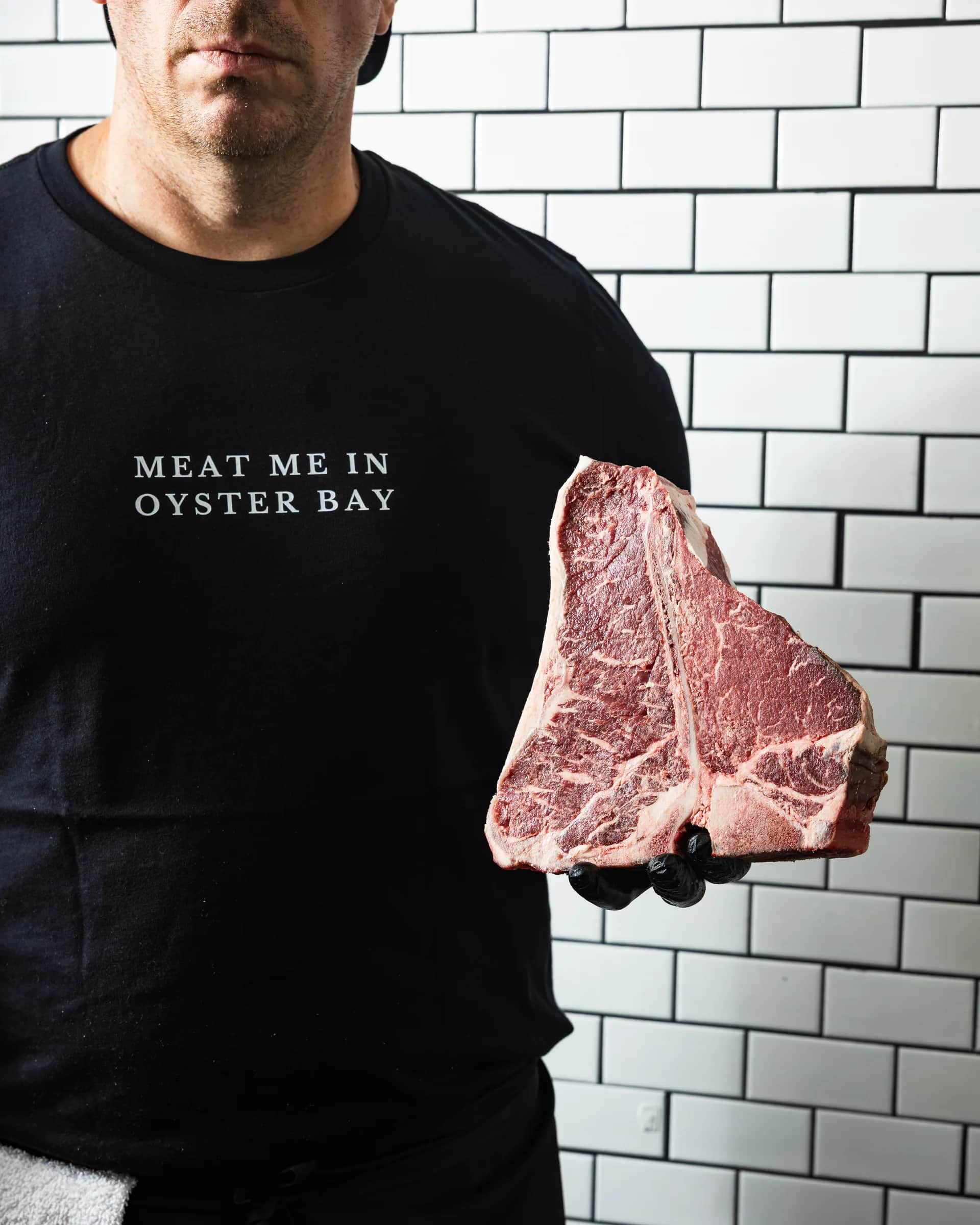 The Bay Butcher tee holding a T-bone against subway tile